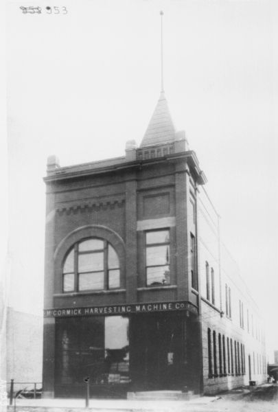 View across street of the exterior of a dealership. Brick building with a large show window on the first floor. The second floor has a large arched window. On the roof is a tower with a flag pole.