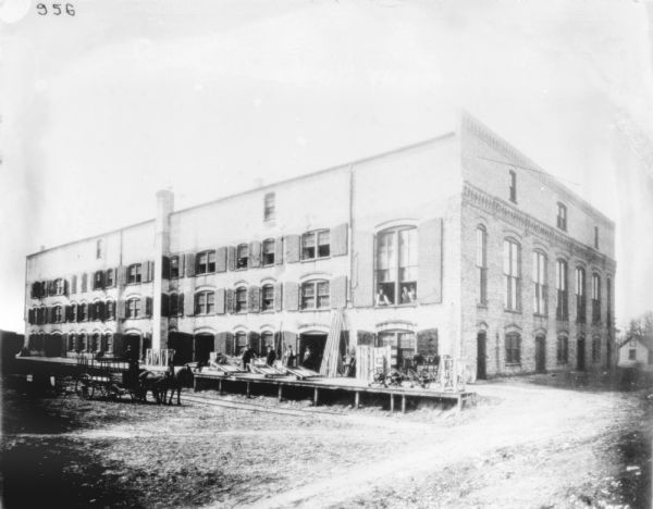 View of group of men posing on and near the loading dock of a dealership. One man is sitting on a horse-drawn wagon on the left, other men are on the loading dock with machinery, and other men are looking out of a window on the second floor on the right. All the windows above the loading dock have wood shutters.