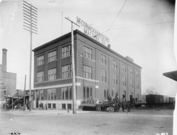 View across street towards men standing in front of the dealership. Two men wearing suits stand on the sidewalk near an entrance on the left. Two man wearing work clothes stand on a horse-drawn wagon backed up to the end of the loading dock. Further down the loading dock are two men standing wearing suits. There are railroad cars on railroad tracks in the background.