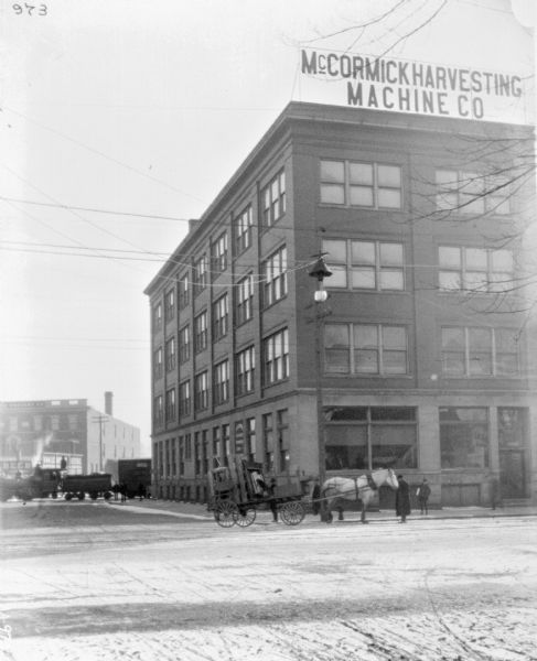 View across street towards the front and left side of a dealership. On the street and sidewalk at the corner men and a boy stand near a horse-drawn wagon. Behind the dealership on the left is a locomotive pulling train cars.