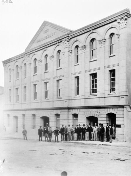Exterior view across street towards a group of about twenty men, many wearing bowler hats, standing in front of the three-story stone building.