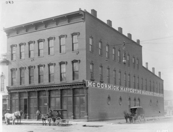 View across street of exterior of McCormick Harvesting Machine Co. dealership. Two men are standing on the sidewalk in front of the three-story building. A man is sitting in a horse-drawn carriage at the curb. There are two other horse-drawn carriages on the left and right.