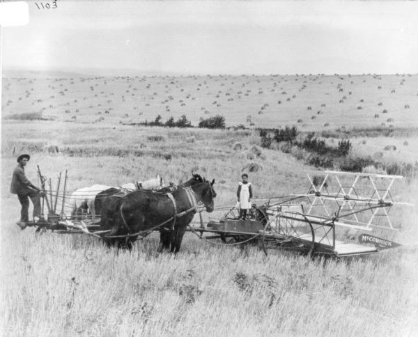 A man is sitting behind a team of horses which is behind a McCormick push binder in a field. A young girl is standing on the push binder.