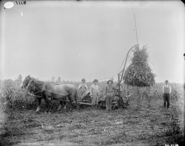 Group of men posing with a horse-drawn corn binder in a field. The machine is lifting the corn shock up in the air.