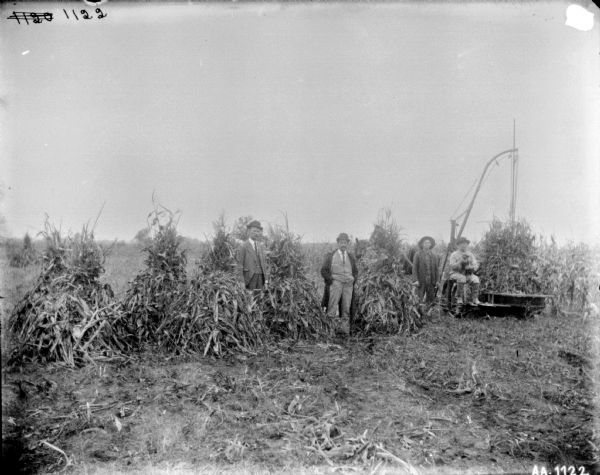 Group of men posing with a corn binder in a field. The man sitting on the corn binder on the right is holding a dog in his lap.
