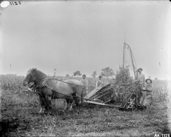 Two men are posing on a horse-drawn corn binder in a field. Two men, wearing vests, neckties, and hats are standing behind the binder.