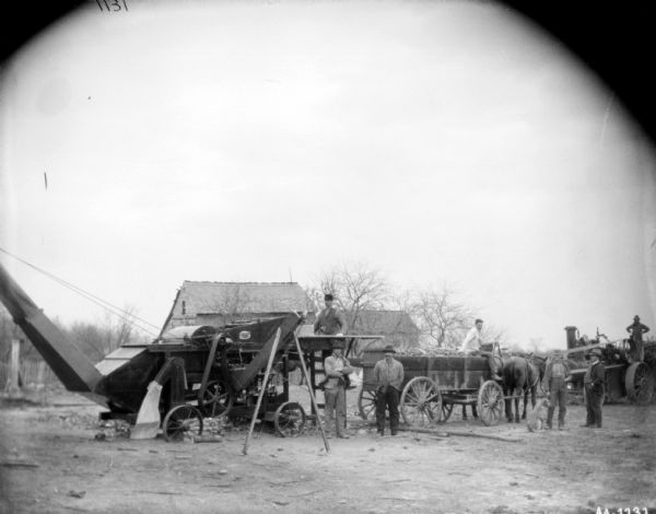 Group portrait of seven men and a dog posing with a tractor powered husker and shredder. The tractor is on the far right powering the belt-driven husker and shredder in the center. A man is sitting on a horse-drawn wagon in the center, which appears to be full of corn cobs. Farm buildings are in the background.