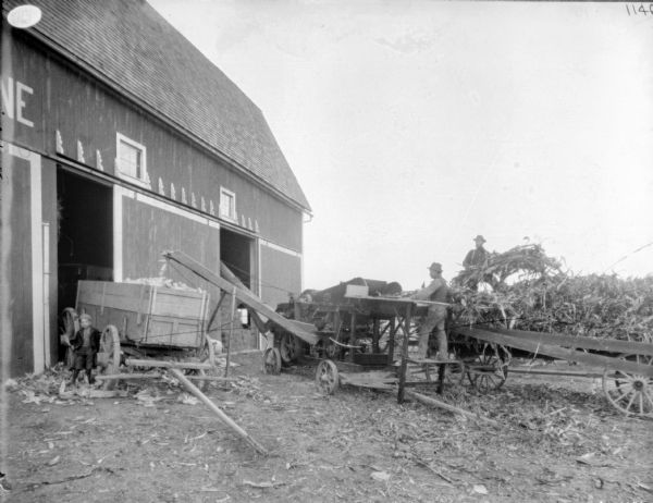 Silaging operation near a barn with two open doors. In the center, two men work unloading a wagon into a belt-driven husker and shredder. A chute is extended into an open barn door on the right side of the barn. There is a young boy posing near a wagon being filled with corn cobs from another chute near the open barn door on the left.