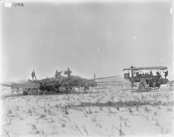 View across snowy field towards men engaged in a silaging operation with a tractor powered husker and shredder. Three men are standing on horse-drawn wagons on the left, and on the right a man is sitting on a tractor.