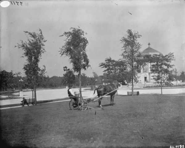 View across well-manicured lawn towards a man using a horse-drawn mower. The horse is wearing a fly-net. Across a drive is a domed pavilion. A man is sitting on one the of the many benches that are along the drive, which is also lined with numerous lampposts.