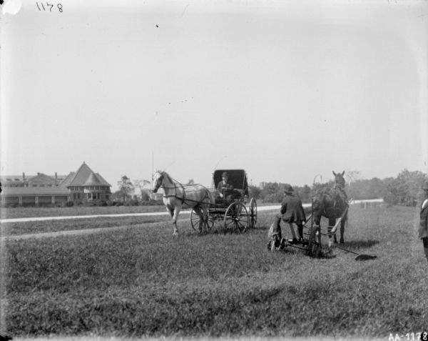 View across well-manicured lawn towards a man sitting on a horse-drawn mower, talking to a man in a horse-drawn buggy. The horses are wearing fly-nets. There is a large building in the background on the left. Another man is standing on the far right.