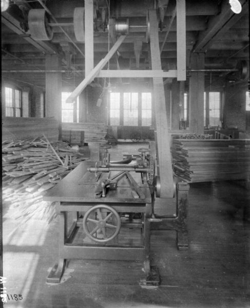 Interior view inside factory of a manufacturing area. In the foreground is a belt-driven machine on a table. On the left is a pile of pieces of loose lumber, and near the windows are stacks of more lumber.