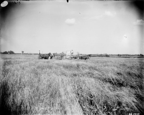 View across field towards men working with a horse-powered push binder header in a field. One man is standing on the push binder. Other men are standing in a horse-drawn wagon near the side of the push binder, and also in the field in the far background.
