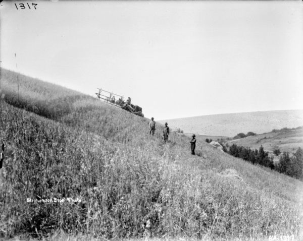 View along side of hill towards three men standing in the field. Behind them further up the hill is a man using a horse-drawn push binder header. A valley and a hill are in the background on the right.