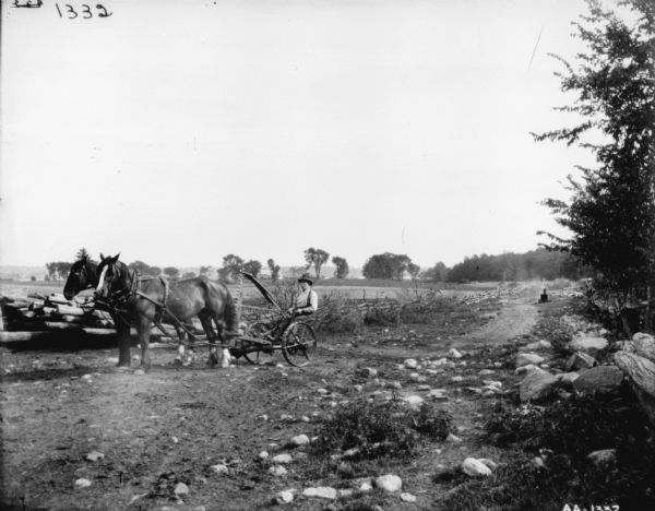 Man using a horse-drawn mower along the edge of a rocky path. There are split-rail fences along the sides of the path. A pile of lumber is behind the team of horses. There are farm buildings among trees in the far background.