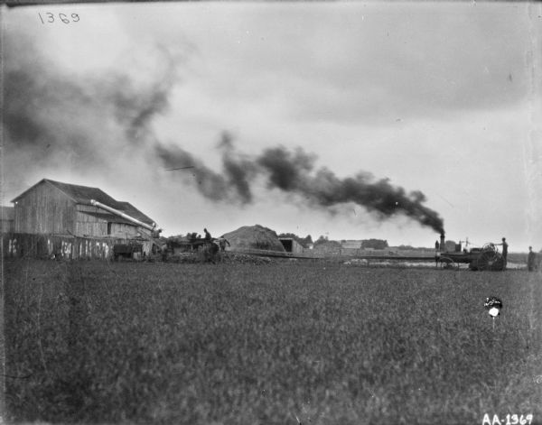 View across field towards men using huskers and shredders near farm buildings. On the right a man is using a tractor for belt-driven power for the husker and shredder that a group of men are using on the left.