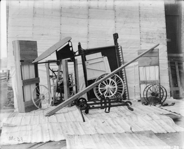 Corn binder dismantled for shipping. Some parts are crated. There is a white sheet behind the corn binder serving as a backdrop, and railroad tracks run along a canal or river in the background on the left.