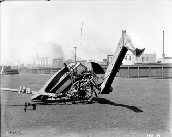 Corn picker set up outdoors. In the background are a fence and industrial buildings.
