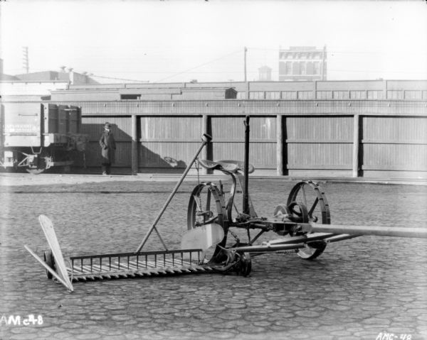Mower with rake and platform sitting on cobblestones outdoors. In the background a man is standing on railroad tracks near a railroad car. Behind the man is a fence and industrial buildings.