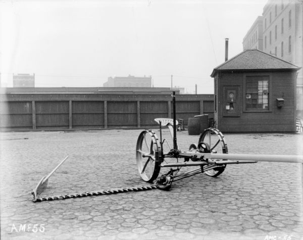 Mower set up on the cobblestones near a factory. In the background is a fence, and a small building, perhaps a guardhouse.