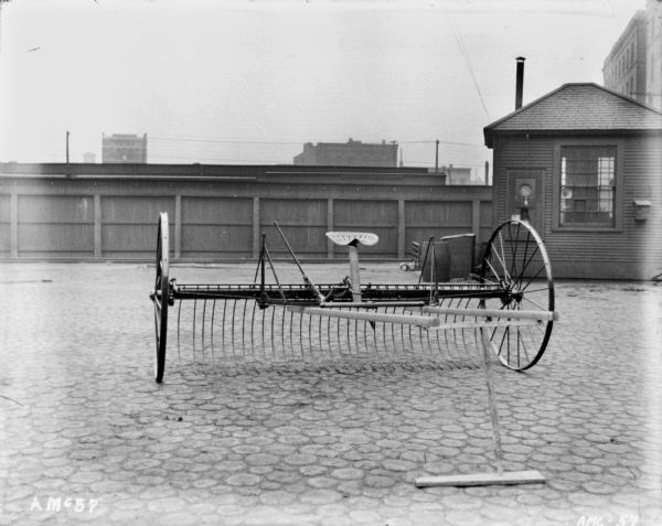 Dump rake set up on the cobblestones near a factory. In the background is a fence, and a small building, perhaps a guardhouse.