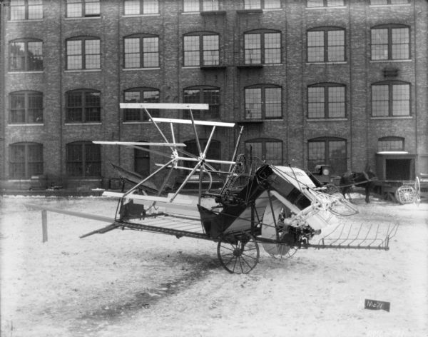 McCormick binder set up on the snow-covered cobblestones in front of a brick factory building. In the background a horse hitched to a wagon stands near a loading dock.