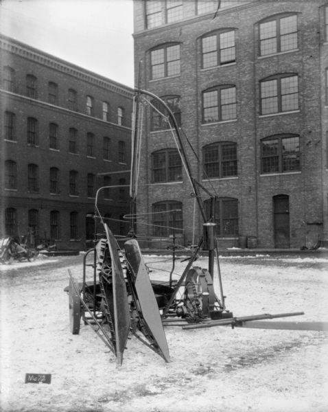 Corn binder, with  attachments and stalkers, set up on snow-covered cobblestones in front of brick factory buildings.