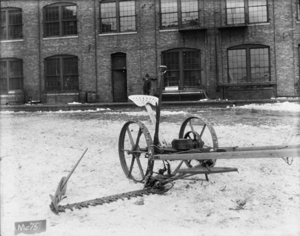 Mower set up on the snow-covered cobblestones in front of a brick factory building. In the background a man is standing on the wooden sidewalk near the doorway of the factory.