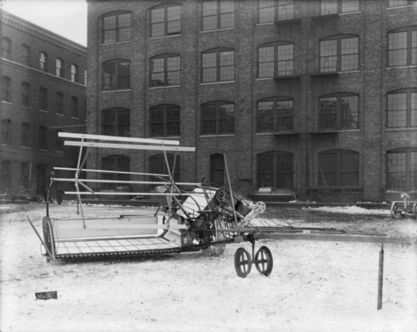 McCormick binder set up on the snow-covered cobblestones in front of a brick factory building. In the background a man is standing between two brick buildings.