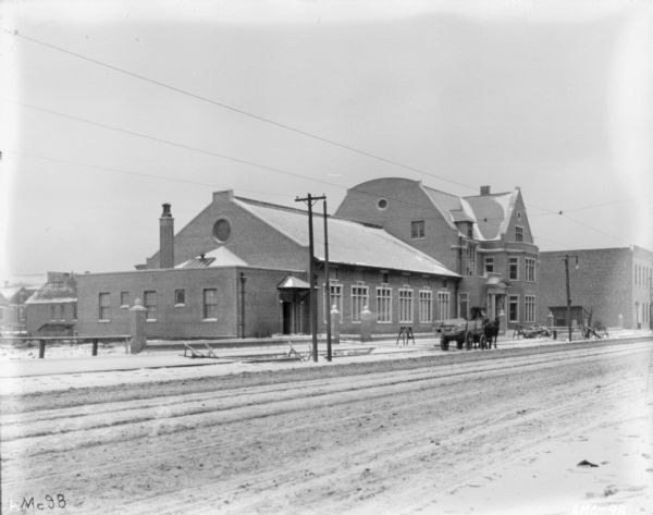 Exterior view across street in winter of the McCormick Works Club House. In front of the club house is a man standing on a loaded horse-drawn wagon parked at the curb near where there is construction of the front walkway.