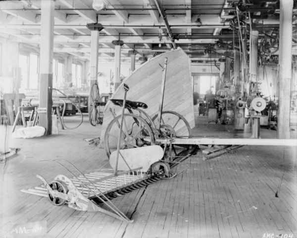 Mower and rake set up on a factory floor at McCormick Works. In the background on the right is belt-driven machinery.
