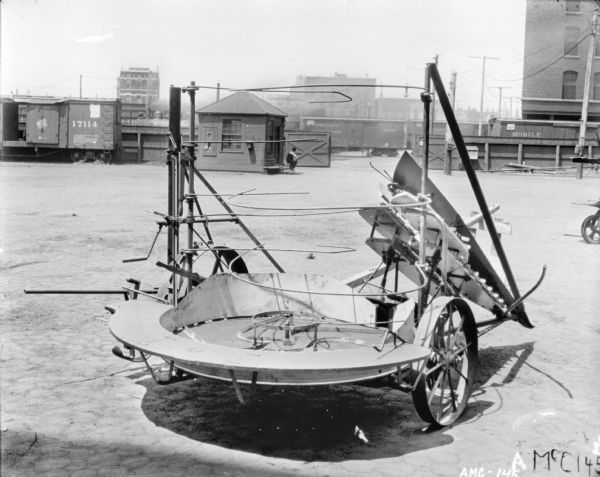 Corn Binder set up outdoors in the yard of McCormick Works. In the background a man is sitting near a small building that my be a guardhouse near railroad cars on tracks and a fence. A factory building is on the right.