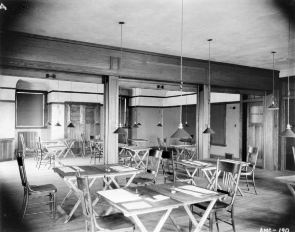 Interior view of the game room at the McCormick Works Club House. Tables and chairs are set up throughout the room. Shades on the windows are drawn. Numerous lamp fixtures are hanging from the ceiling.