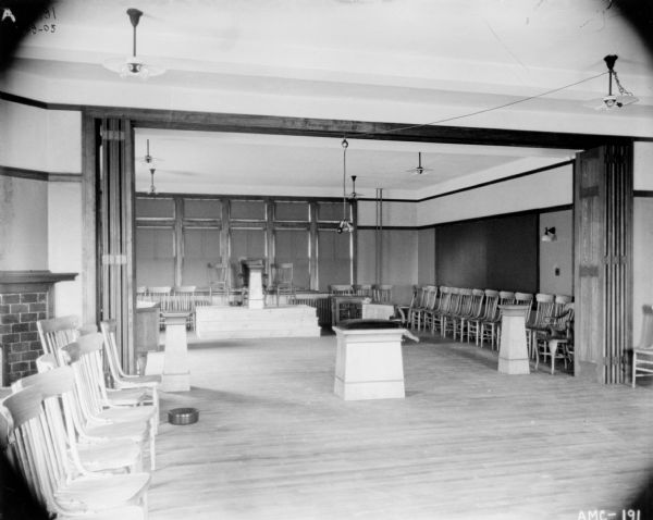 Interior view of lecture room. Chairs are in rows along the walls. There are chairs on a raised platform at the back of the room just in front of windows with the shades drawn. A spittoon is on the floor near the folding wall divider on the left.