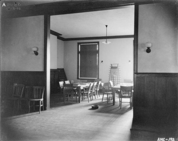 Interior view, from another room or hallway, of the meeting room. Chairs, tables, and what is probably a newspaper rack are in the room. There is a spittoon on the floor at the entrance to the room. The shade on the window is drawn.