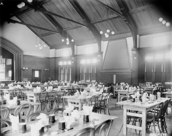 Interior view of the dining room. Tables and chairs are set up in the large room which has a vaulted ceiling, with chandeliers hanging from the cross braces. The tables are set with mugs, silverware, and napkins in glasses. A large brick fireplace is along the side wall flanked by tall windows, with some of the shades drawn. A brick archway is at the end of the room on the left, and may be framing a stage.