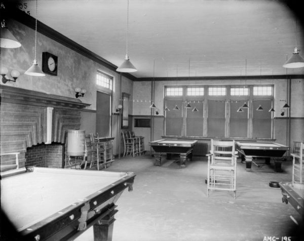 Interior view of the Pool Hall. There is a brick fireplace along the wall on the left. Chairs are set along the walls. Two pool tables are in the foreground, and two more are in the background. The shades on the windows are drawn. Numerous lamps are hanging from the ceiling above the pool tables. Spittoons are on the floor.