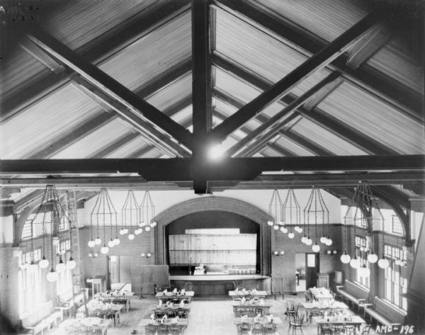 Elevated view of the dining room. The vaulted ceiling has chandeliers hanging from the cross braces. Below, tables and chairs are set up in rows, and benches are set against the windows on both sides of the room. In the front of the room is a stage with a brick arch. Numbered panels of wood and other materials are on the stage. Part of a large fireplace is on the far right.