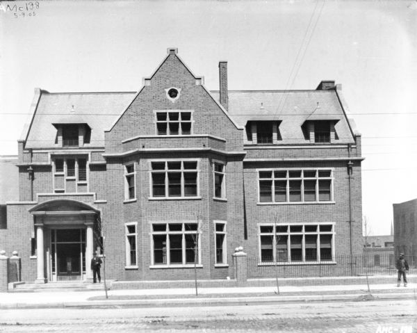 View across street of the exterior of the McCormick Works Club House. A man is standing near the entrance, and another man is standing on the sidewalk on the right.