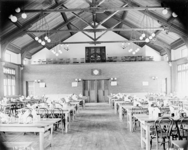 View of the back of the dining room towards an open balcony above a brick wall. There are rows of chairs set up on the balcony. The dining room has a vaulted ceiling with chandeliers hanging from the cross braces. Chairs are set up at the dining tables.