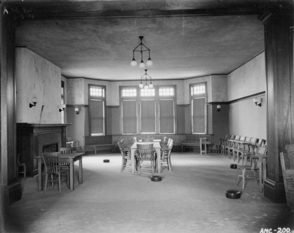 Interior view of the meeting room, On the left is a fireplace. Tables and chairs are in the center of the room. At the back of the room is a bay window with benches. On the right along the wall is a row of chairs. A number of spittoons are on the distributed about on the floor.