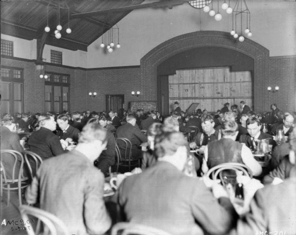 View of mostly male, and a few female employees, dining in the Dining Hall of the McCormick Works Club House. In the background is the stage. Numbered wood panels close off the front of the stage. To the left of the stage near a doorway is a blackboard with the "Bill of Fare" written in chalk.