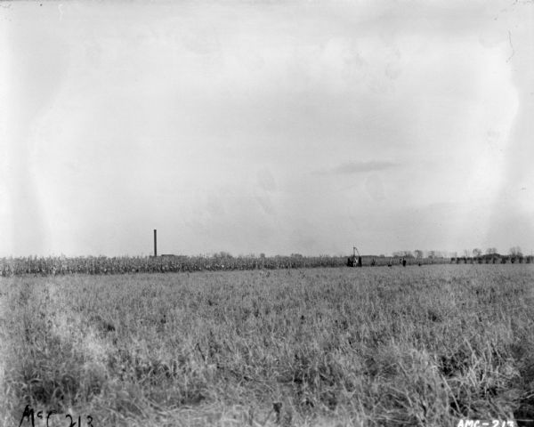 View across field towards men standing near a man using a corn binder in a field to bind stalks.