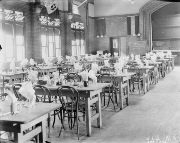 Interior view of dining room. Rows of tables are set with napkins in drinking glasses and mugs. Flags are hanging over the windows on the left and along the back brick wall. The edge of the arched stage at the front of the room is on the right.