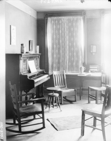 Interior view of the music room. There is a piano with a piano stool against the wall on the left. Two rocking chairs, two chairs and a desk are gathered around the piano in front of two windows with drawn shades.