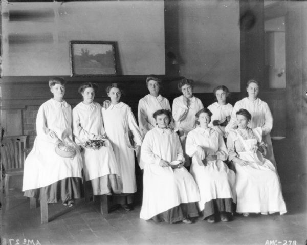 Group portrait of women wearing long white smocks posing with kitchen utensils, baskets, cups, plates and flowers. The women are sitting on a table and chairs. There is a large fireplace with a painting resting on a mantel behind them. They may be posing in the McCormick Works Club House.