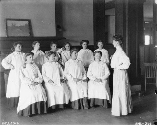 Women dressed in white smocks posing with kitchen utensils. The women are posing sitting and standing. A woman wearing a long skirt and a blouse is standing in profile on the right facing them. There is a large fireplace with a painting resting on a mantel behind them. They may be posing in the McCormick Works Club House.