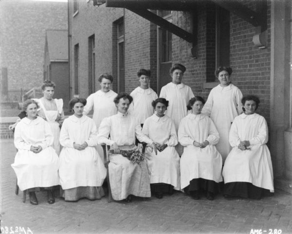 Group of women sitting and standing outdoors, perhaps at the McCormick Works Club House. All the women are wearing long smocks over dresses, except the woman sitting front and center, who has a bouquet of flowers on her lap.