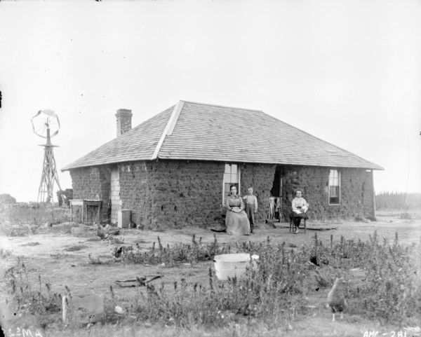 View of farm woman and her two children posing in front of sod farmhouse on poor plot of land. The woman is sitting in a chair near the open front door. A young boy is standing beside her, and a young girl is sitting in a chair holding a doll. Chickens are roaming in the yard, and in the background on the left behind the house is a windmill.