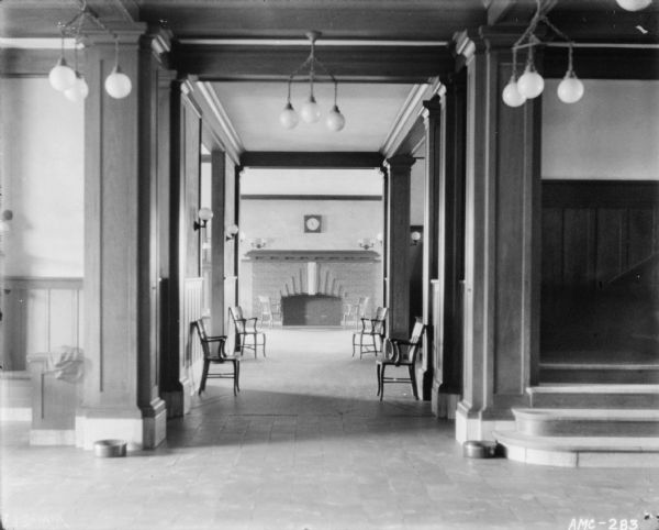 View from tiled lobby towards an interior hallway leading to a room with a brick fireplace. On the right of the lobby is a staircase, and on the left are built-in benches. Spittoons are on the floor near the benches and the base of the stairs.